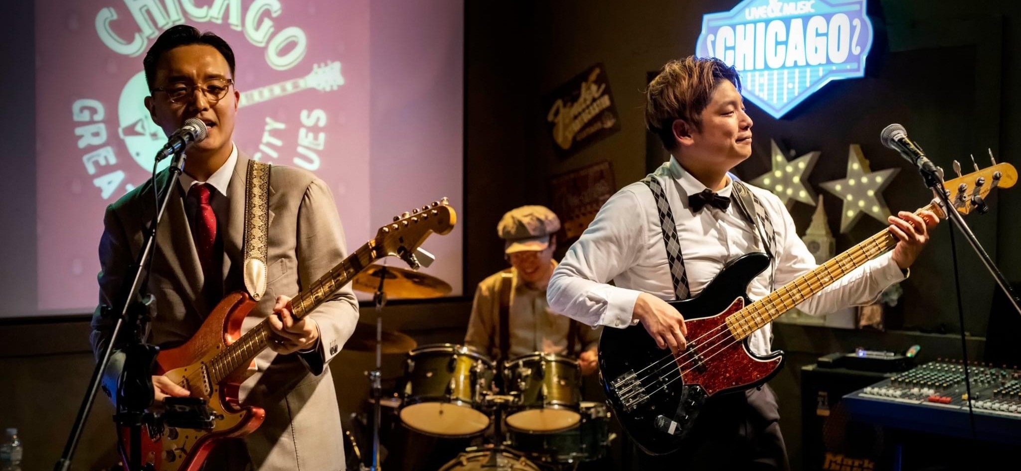 Three men playing instruments, one with a guitar, singing, one with a bass guitar, and behind them, one playing the drums.